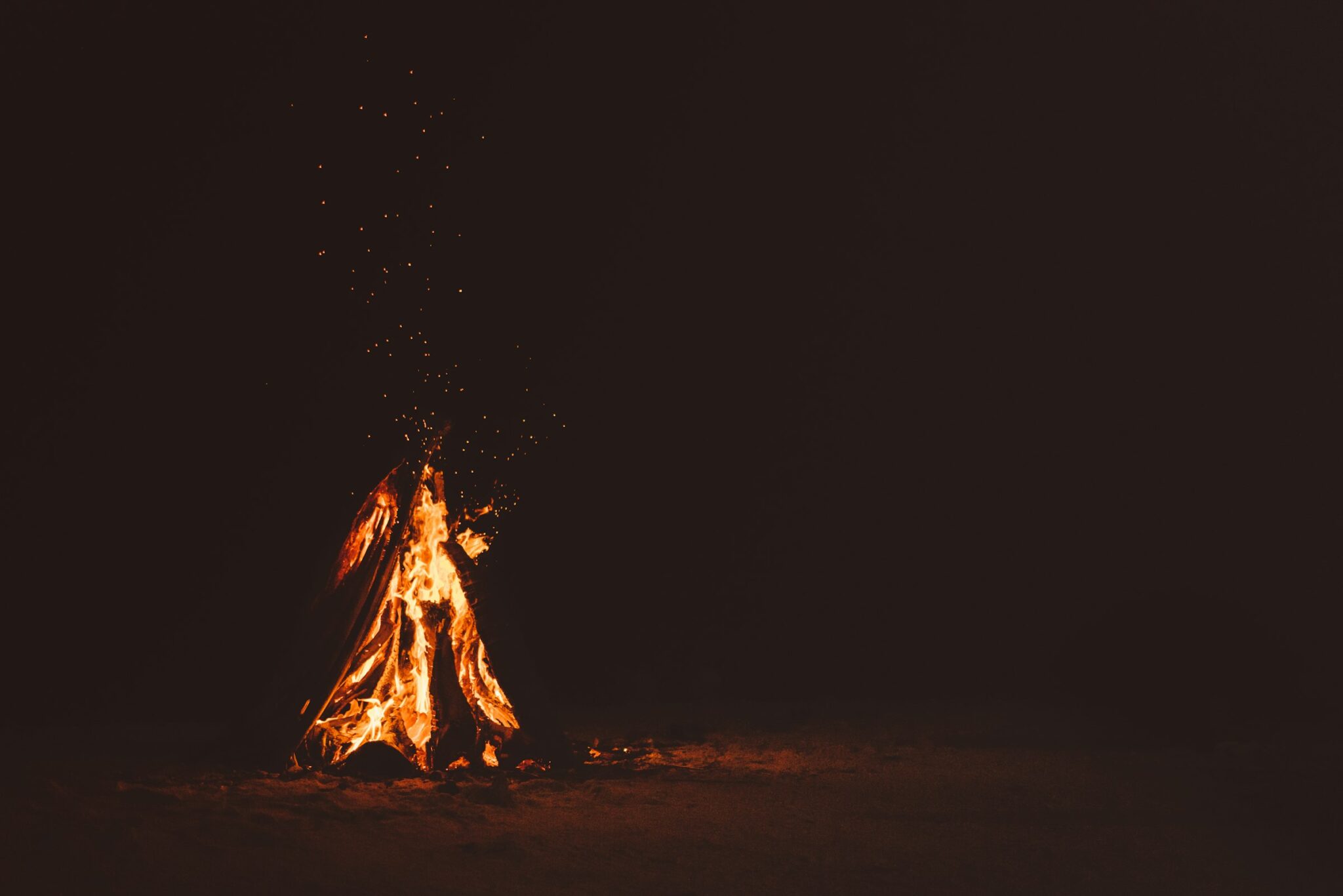 A single fire burning on dark sand at night, sparks rising